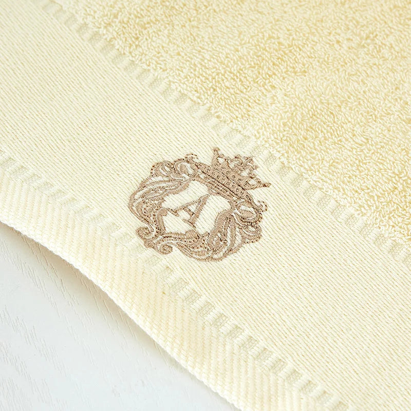 Cream cotton towel with embroidered gold crest on the corner and stitched border edge.