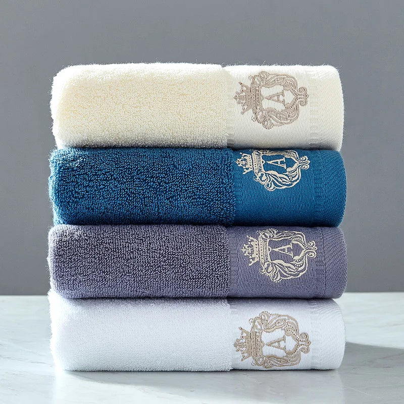 Embroidered cotton towel set with folded white, blue, and dark gray towels, gold crest trim.