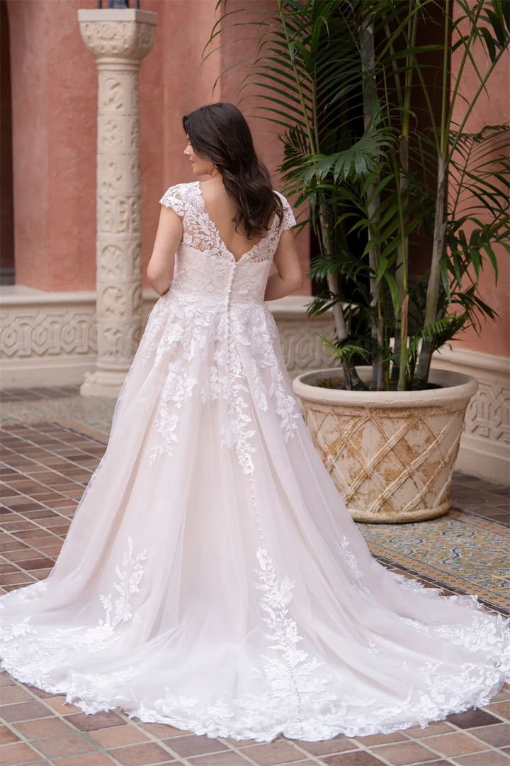 Woman in a white lace wedding dress standing in an outdoor setting with plants and a building in the background.