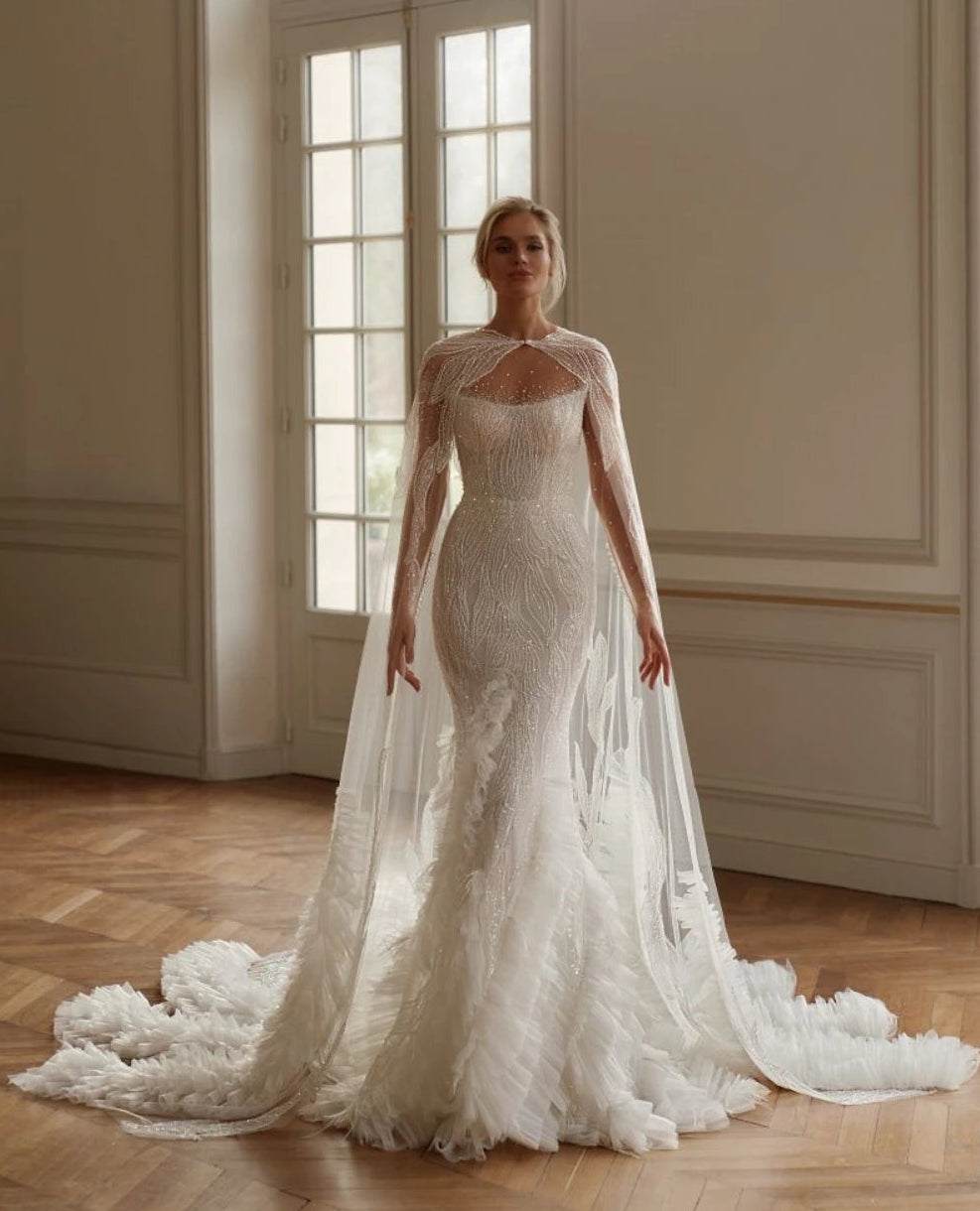 Woman in a white lace wedding dress standing in a room with large windows.