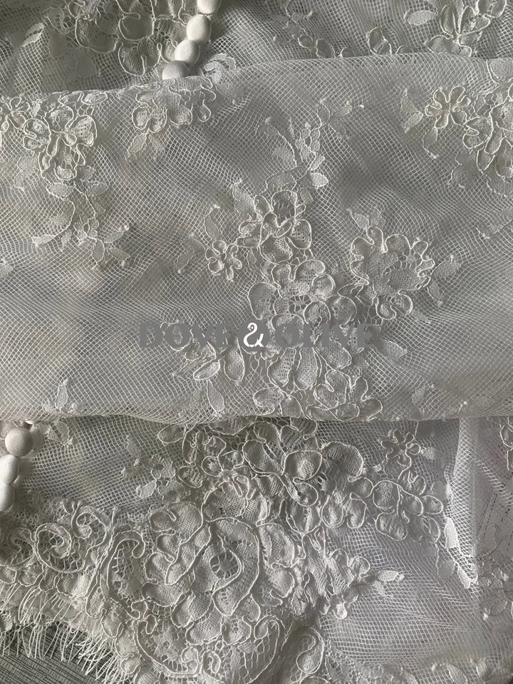 Off-white lace tulle fabric with dense floral embroidery and scalloped edge