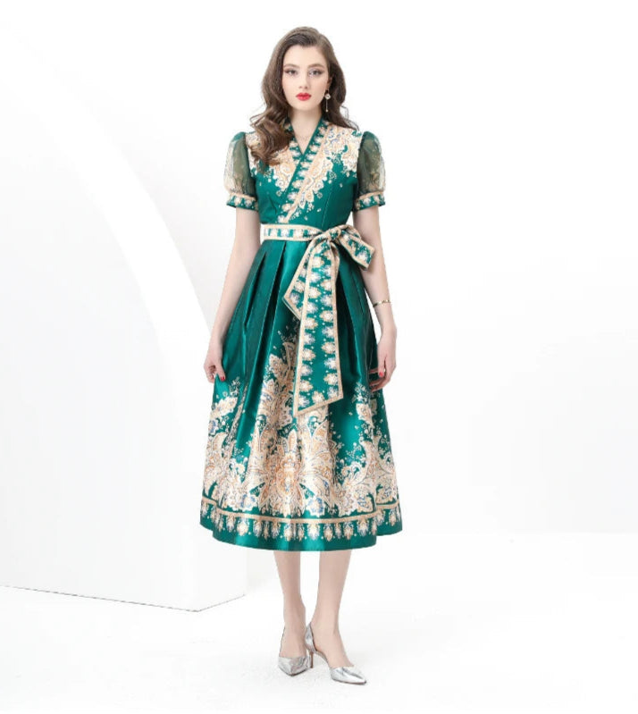 A model wearing a green floral print satin wrap midi dress with short sleeves and a V-neckline, waist tie, and mid-calf length.