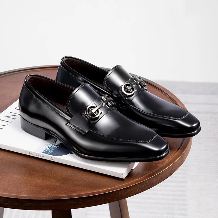 Black leather slip-on loafers with shiny buckle detail on a wooden display