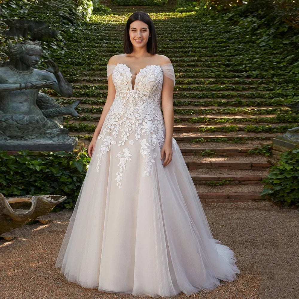 A woman standing in a garden wearing a white, off-the-shoulder, sweetheart neckline, sleeveless, floor-length wedding dress with applique detailing.