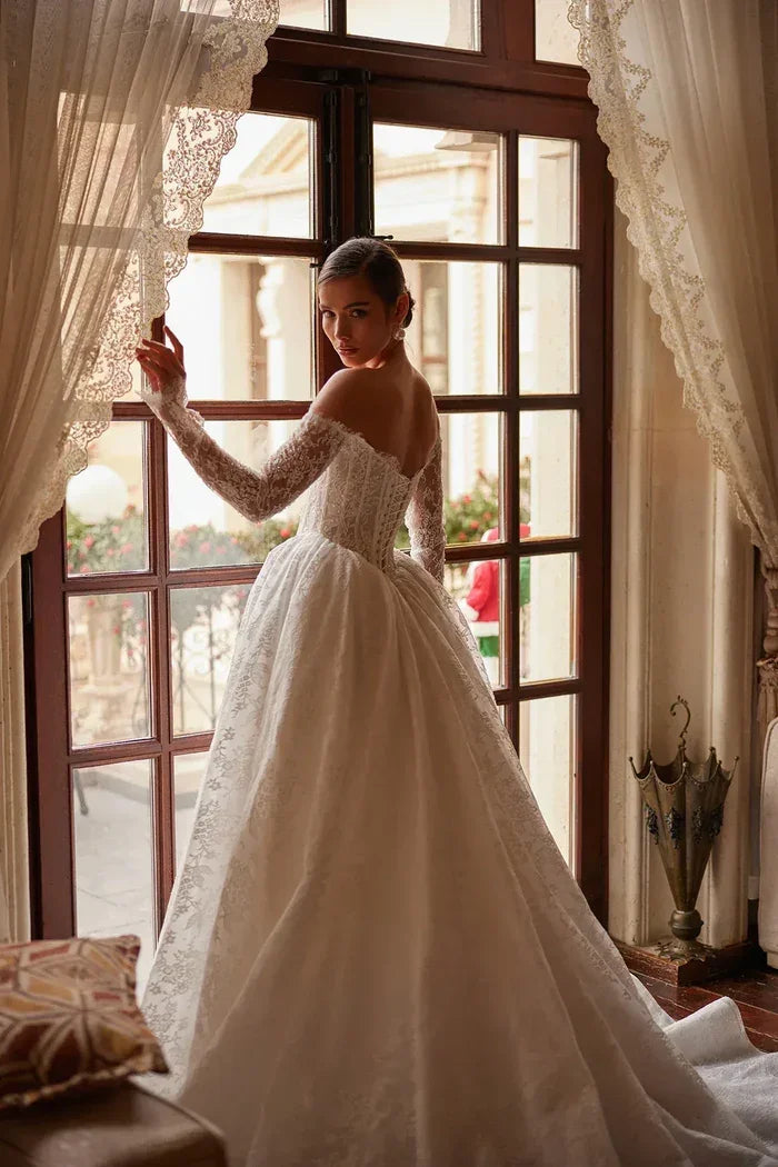 Lace wedding gown with off-shoulder neckline and gathered A-line skirt by window