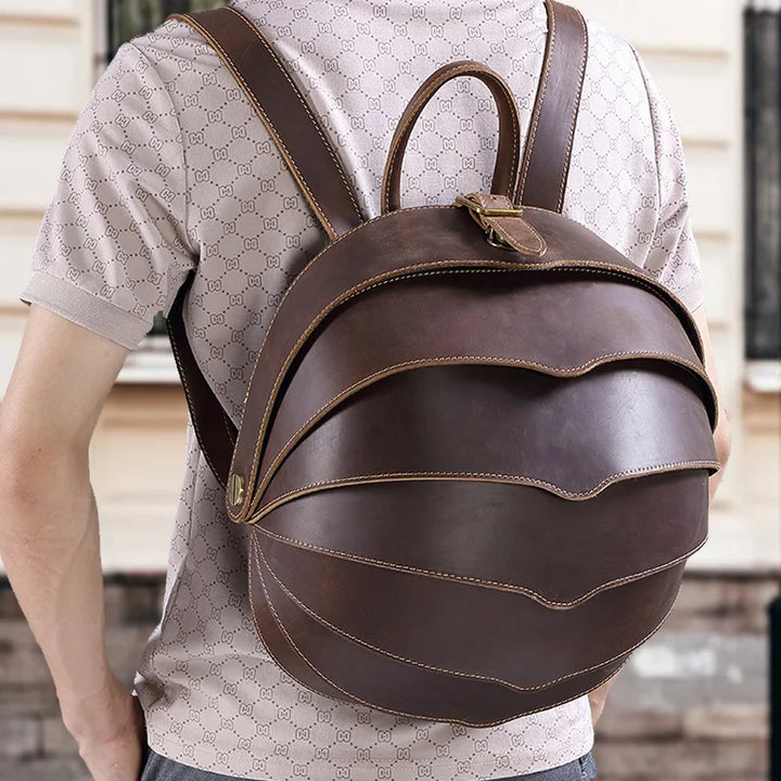 Brown leather round backpack with curved stitched flap and top handle; worn on model.