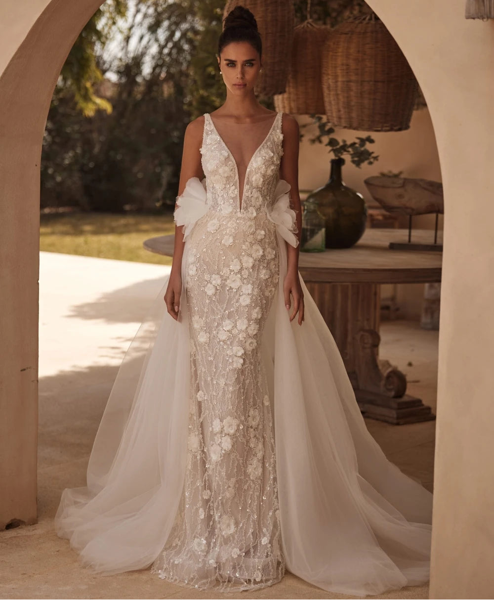 Woman in a white floral wedding dress standing in a rustic outdoor setting with 'YIWANNA' branding.