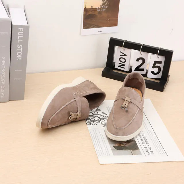 Beige suede slip-on loafers with metal buckle strap and white rubber sole.
