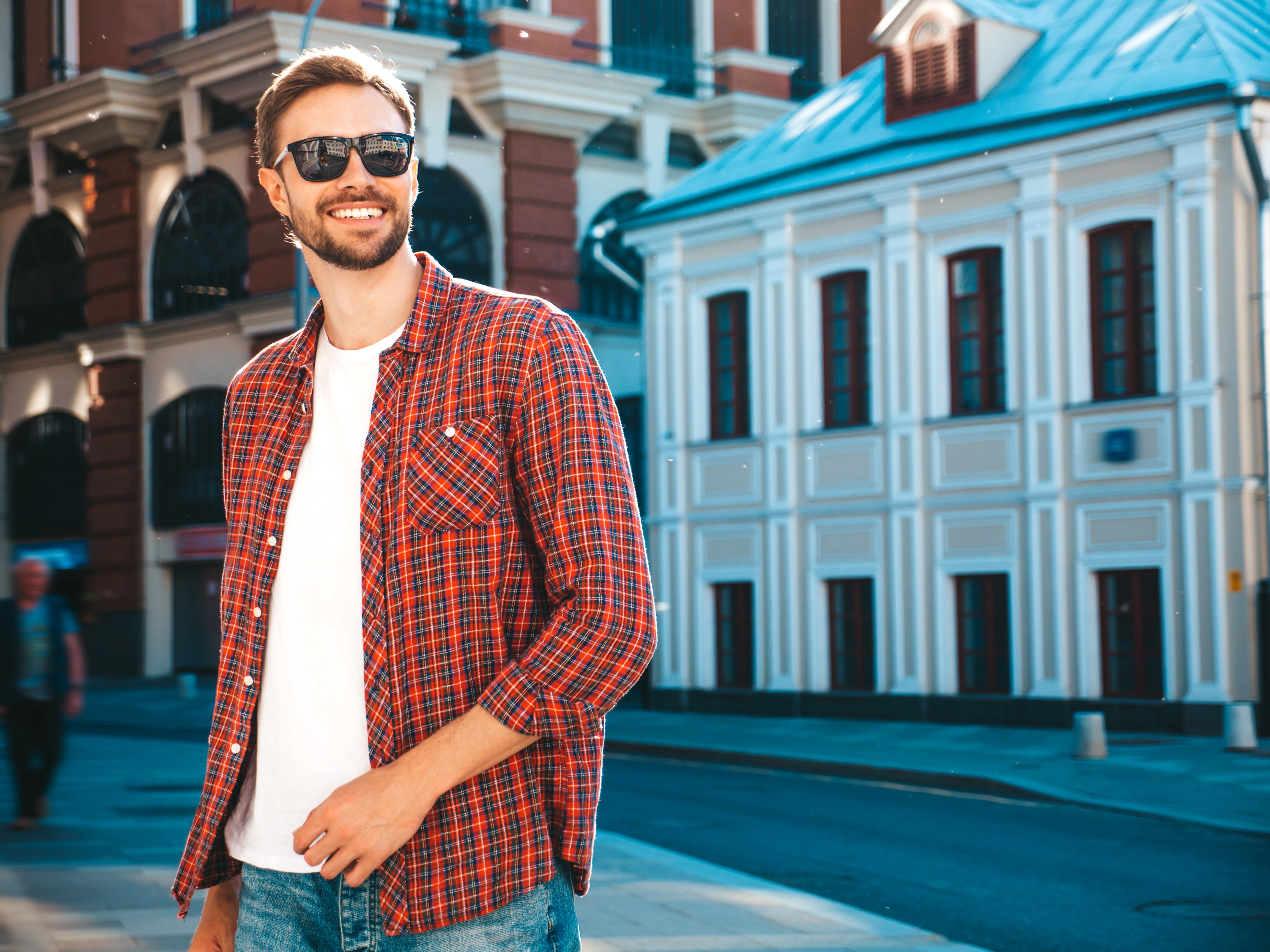 Man wearing sunglasses and a red plaid shirt standing on a street with buildings in the background